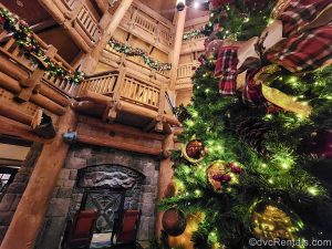 A green Christmas tree adorned with gold and red wilderness-themed ornaments is see in the lobby of the Boulder Ridge Villas at Disney’s Wilderness Lodge. Matching garlands are hung on the upper, wooden railings, and a fireplace with 2 chairs is seen in the background.