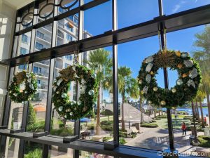 Three wreaths with gold and silver ribbons and baubles are displayed on a large window at Disney’s Riviera Resort. Out the window, the resort grounds are seen under a sunny sky