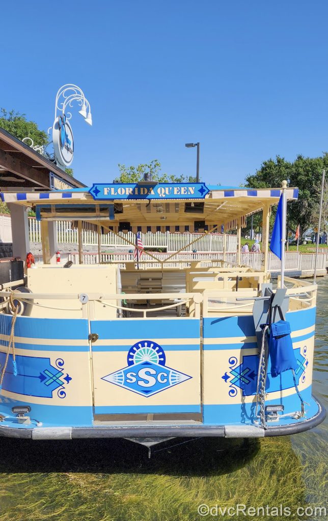 The “Florida Queen” yellow and blue water taxi sits docked at Disney’s Saratoga Springs Resort & Spa under a blue sky, waiting for guests to board.