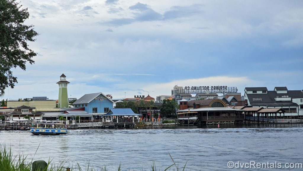 A yellow and blue Disney water taxi sails past the Boathouse restaurant and other buildings at Disney Springs under a cloudy sky.