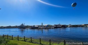 Paddlefish and other restaurant and shop exteriors as well as the Aerophile balloon attraction at Disney Springs are seen from across the water under a sunny sky from Disney’s Saratoga Springs Resort & Spa.