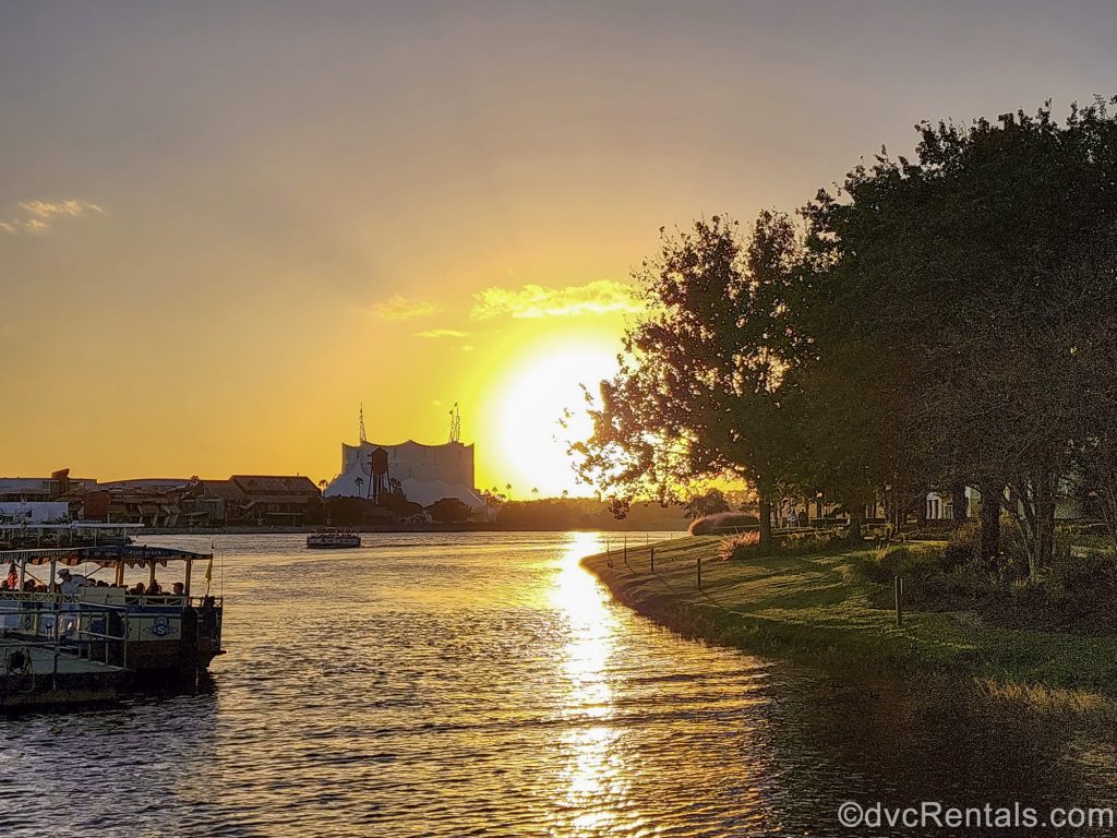 Water taxis sail between Disney Springs and the nearby DVC resorts, Saratoga Springs and Old Key West during golden hour as the sun shines bright, warm yellow light in the distance under a hazy sky.
