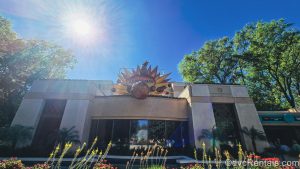 The beige exterior of The Dino Institute is seen under a bright, sunny sky. There is a fireball-shaped sign reading “Dinosaur” atop the building and red and yellow flowers are seen in front of the building.