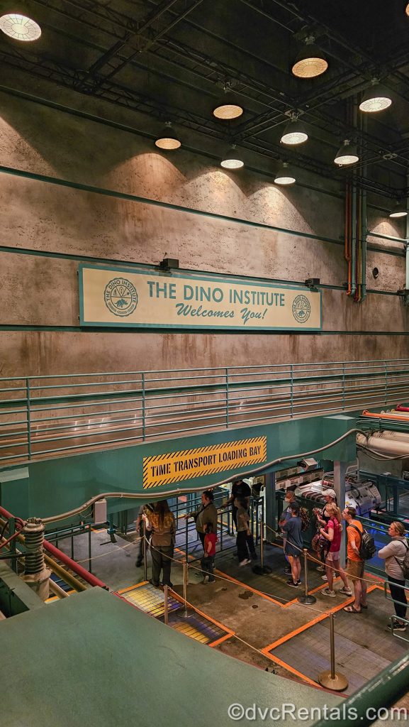 Guests wait to board their ride vehicles in a lower, brick room designed to look like a testing facility. A sign on the wall reads “The Dino Institute Welcomes You!” and another, lower sign reads “Time Transport Loading Bay” below.