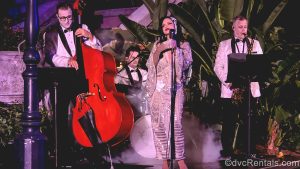 A trio of musicians and a singer all dressed in glamorous formal outfits perform live outside the Tower of Terror attraction as part of the Twilight Soiree at the Tip Top Club during Disney Jollywood Nights.