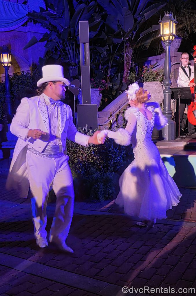 Dancers perform during the Twilight Soiree at the Tip Top Club during Disney Jollywood Nights. Both are dressed in sparkling, white, retro-designed costumes; one a tuxedo and the other a gown and feather-trimmed elbow gloves.