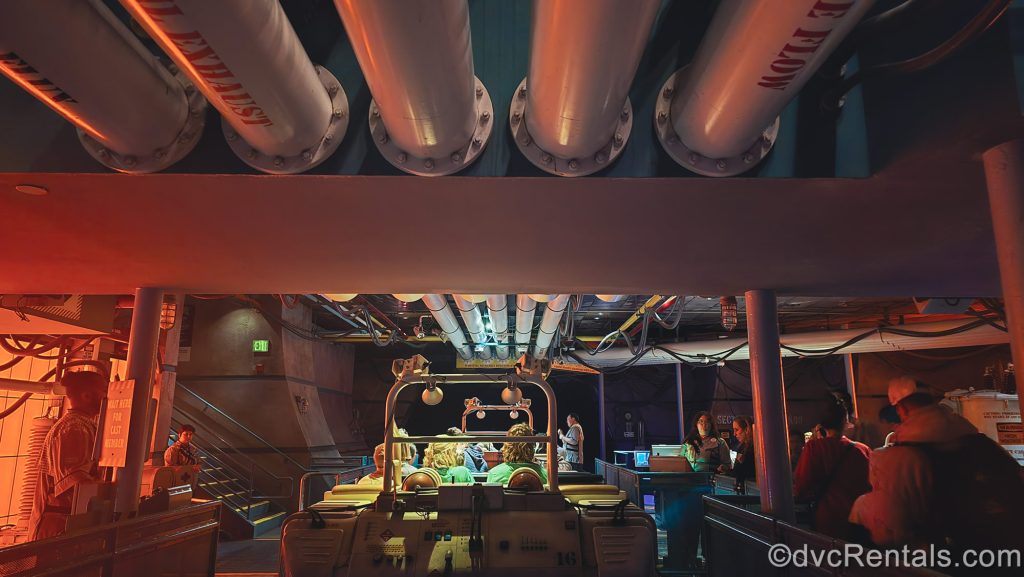 Guests wait to deboard their Time Rover ride vehicles after riding DINOSAUR. The room has many pipes and wires and is illuminated with red and orange lights to make it look like a scientific testing facility.