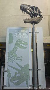 A fossilized dinosaur skull is fixed on top of a pole holding a light blue and green sign for The Dino Institute Gift Shop, which also has drawings of dinosaurs on it.