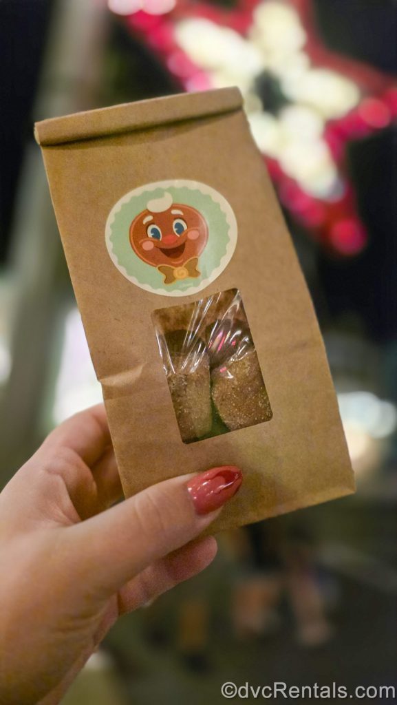 A hand holds up a brown bag full of plant-based Gingerbread Bites. The bag is decorated with a sticker of Ollie the jolly gingerbread man.