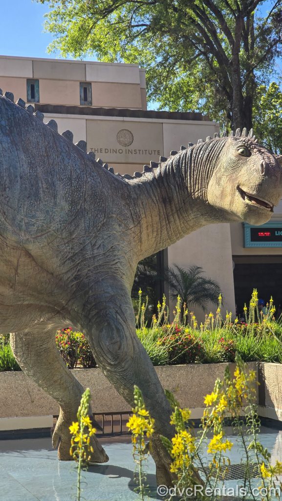A large dinosaur statue is seen outside the DINOSAUR ride building, with green foliage and yellow flowers surrounding it.