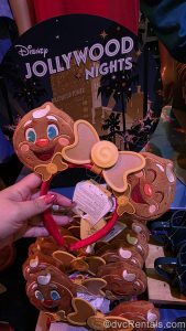 A hand holds a pair of Minnie ears in front of a sign for Disney Jollywood Nights. Each ear is designed to look like the party’s mascot, Ollie the jolly gingerbread man, with a candy-inspired brown bow in the center.