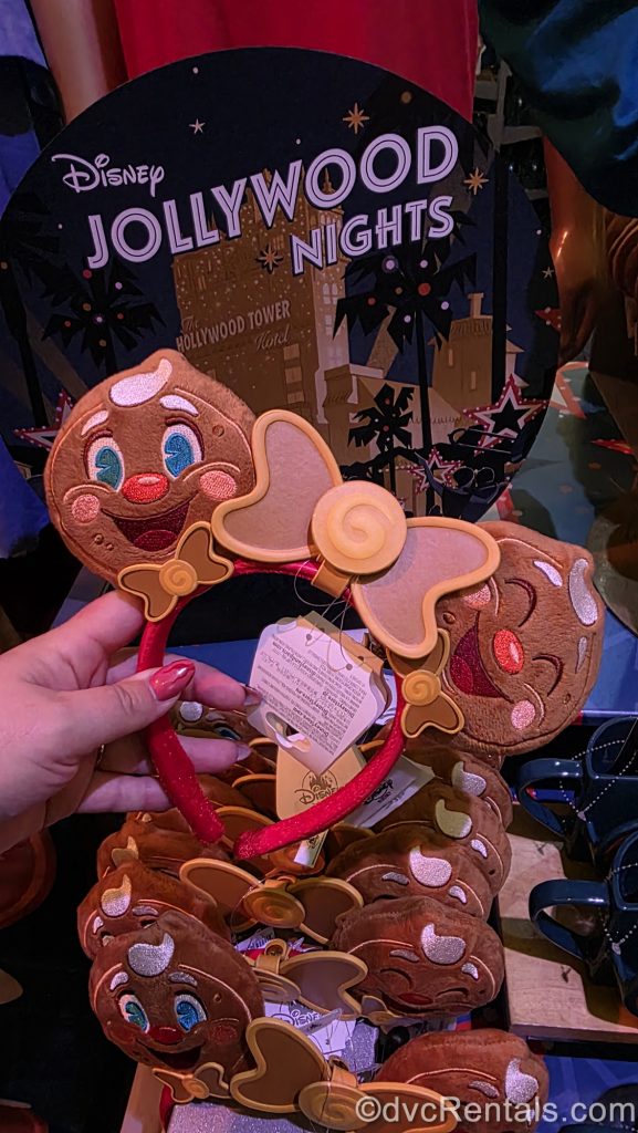 A hand holds a pair of Minnie ears in front of a sign for Disney Jollywood Nights. Each ear is designed to look like the party’s mascot, Ollie the jolly gingerbread man, with a candy-inspired brown bow in the center.