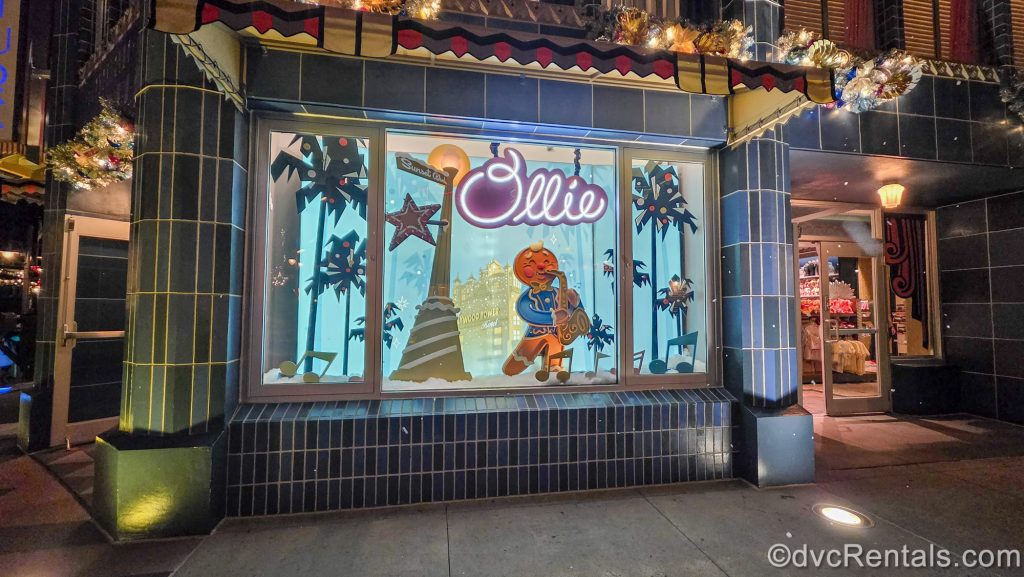 Ollie the jolly gingerbread man is seen wearing his blue jacket playing a gingerbread saxophone under some palm trees in a store window display at Disney’s Hollywood Studios.