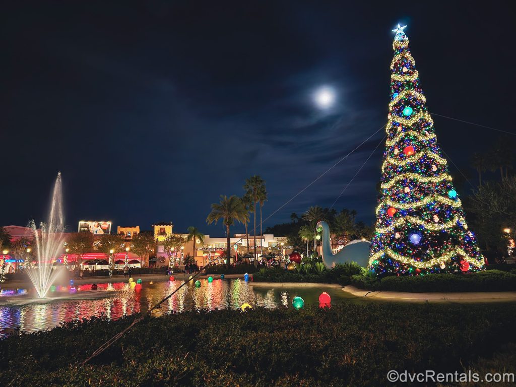 A tall Christmas tree with colorful decorations and lights is displayed next to a water fountain and feature at Disney’s Hollywood Studios under a clear night sky.