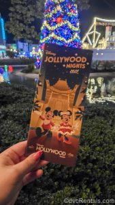 A hand holds up a map pamphlet for Disney Jollywood Nights in front of a brightly lit Christmas tree. The front of the pamphlet features Mickey and Minnie Mouse in seasonal outfits.
