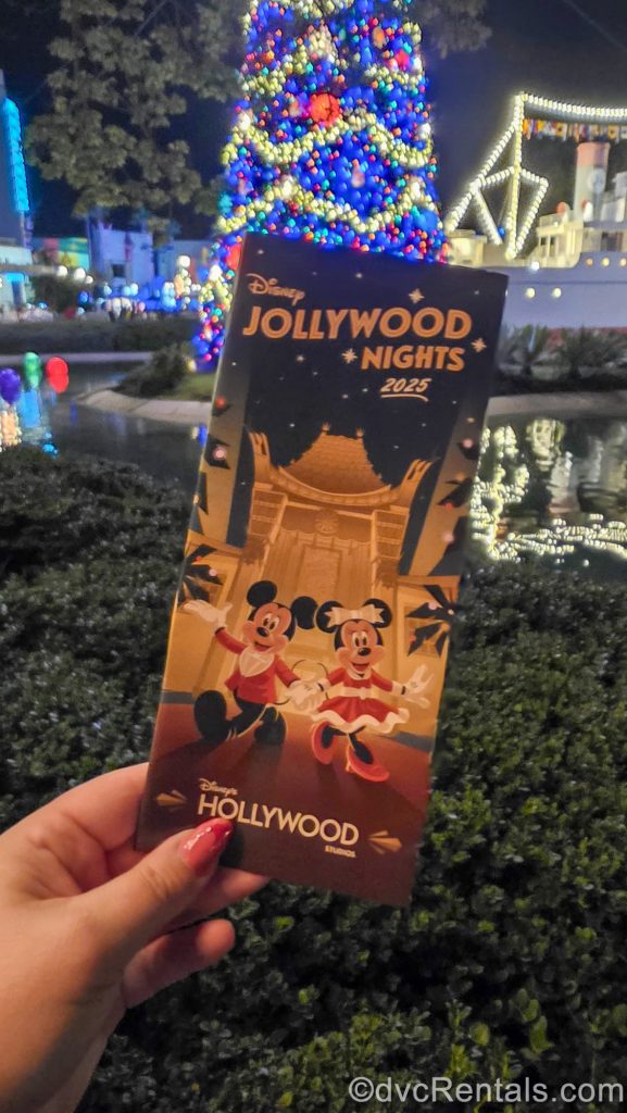 A hand holds up a map pamphlet for Disney Jollywood Nights in front of a brightly lit Christmas tree. The front of the pamphlet features Mickey and Minnie Mouse in seasonal outfits.