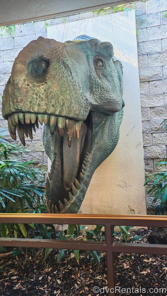 A large, stone sculpture of a dinosaur head with its mouth open and large teeth visible is seen displayed on a brick wall amongst green foliage in the queue for DINOSAUR.