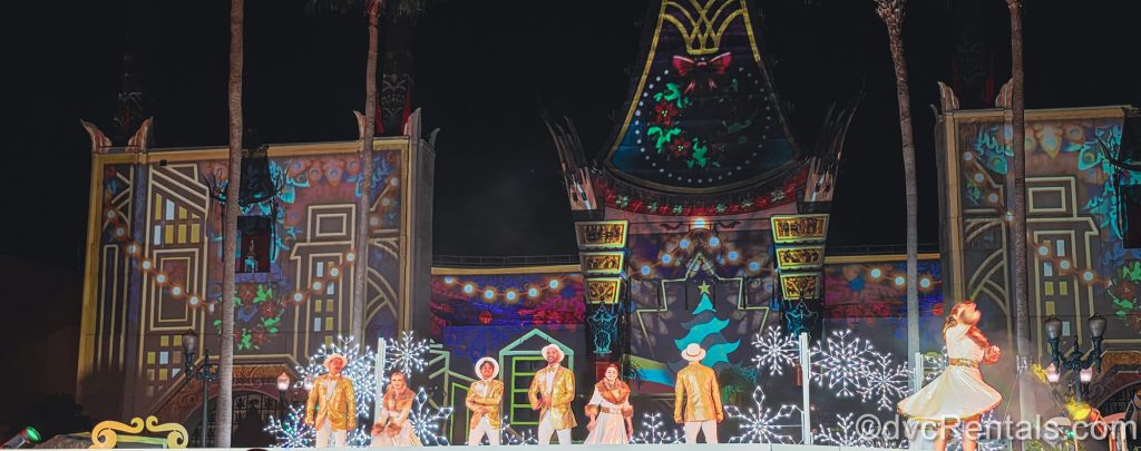Skaters dressed in gold and white holiday season costumes perform in front of the Chinese Theatre during Glisten! The set and Chinese Theatre in the background are decorated with colorful Christmas imagery.