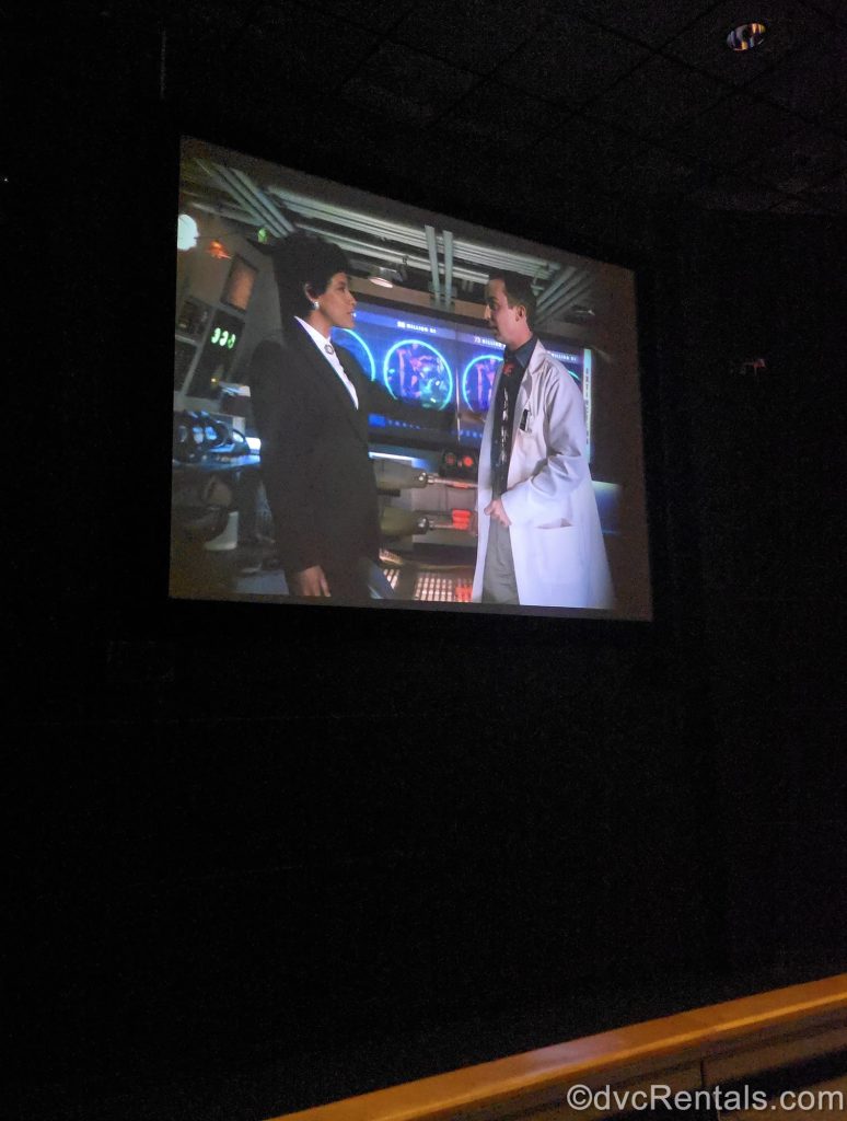 A large screen displays a video of a two actors playing scientists in a computer lab discussing safety and dinosaur information. This video is part of the DINOSAUR pre-show.