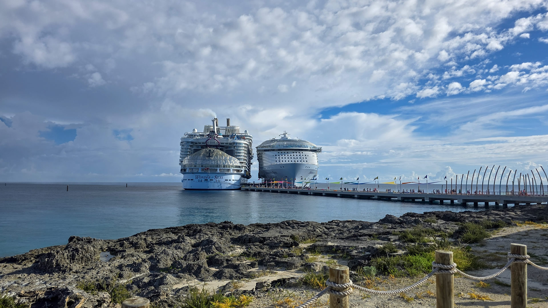 Royal Caribbean’s Wonder of the Seas and Symphony of the Seas cruise ships are docked next to each other at Perfect Day at CocoCay under a sunny sky as guests walk down the pier to enjoy the private island.