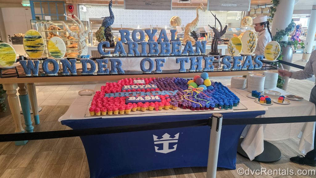 A large display of colorful cupcakes is seen in the Windjammer buffet. Above the cupcakes there is a blue sign reading “Royal Caribbean Wonder of the Seas”, as crew members prepare the desserts for guests.