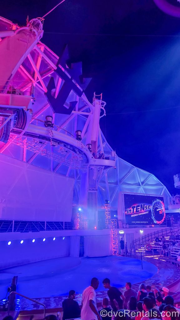 Guests take their seats at the AquaTheater ahead of an inTENse show performance using the pool and diving platforms. Signage for the show is displayed in the venue on a screen to the right under the open-air night sky.