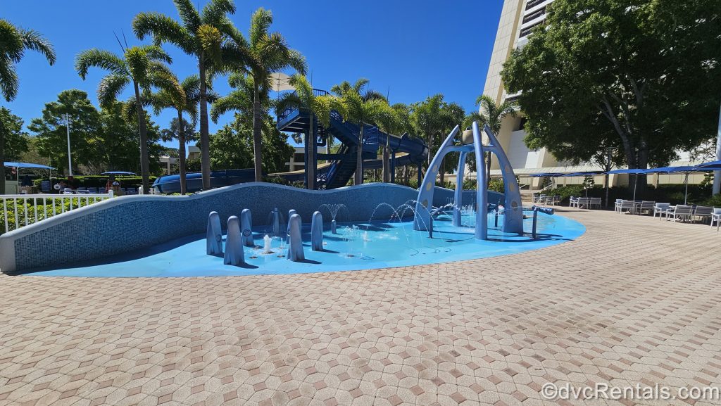 A blue splash pad area with bubbling jets and fountains is seen under a sunny sky and palm trees at Bay Lake Tower.