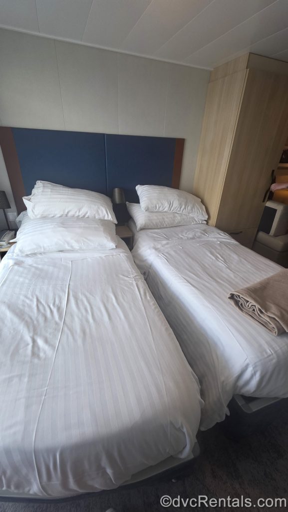 Two twin beds, both with solid white bedding are seen in a stateroom onboard Wonder of the Seas. There is also a wooden storage cupboard next to the beds on the right.