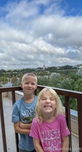 Two of the children in Guest Blogger ErinAdvocates’ family pose on a balcony at Bay Lake Tower with Cinderella Castle in the distance under a blue sky.