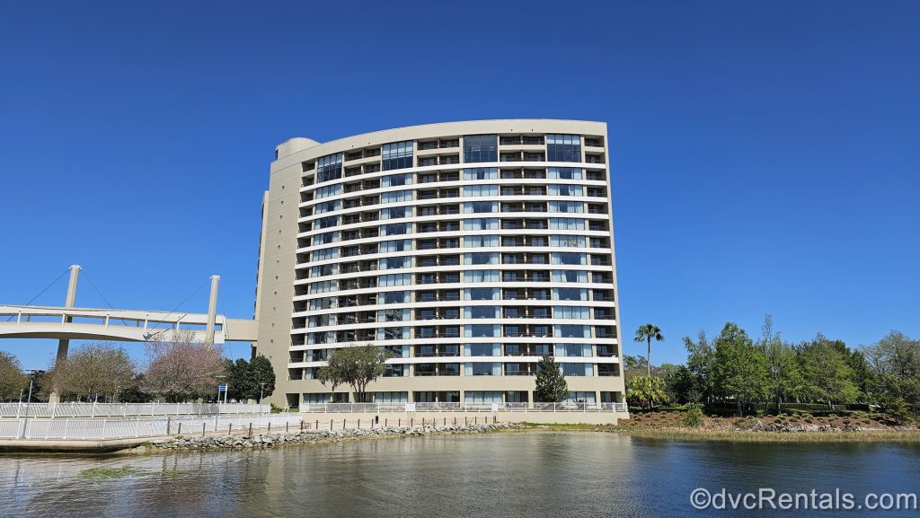 The exterior of Bay Lake Tower and the Sky Way Bridge are seen from across the water under a bright, blue sky.