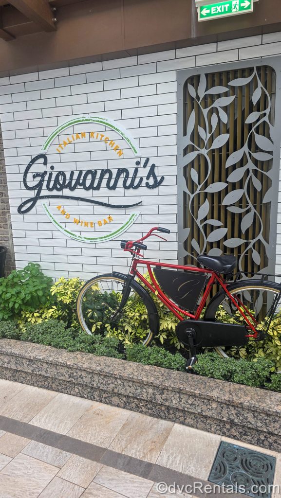 A red bicycle is parked in some green foliage front of a white-tiled wall with the sign for Giovanni’s Italian Kitchen and Wine Bar.