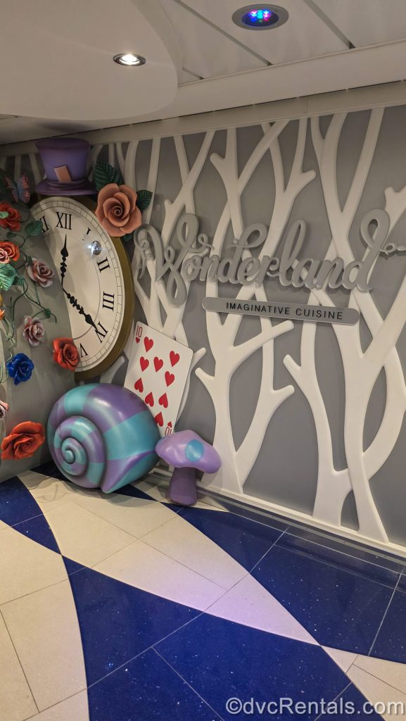 A overly large clockface with a purple hat on top, along with oversized colorful flowers, a playing card, and other Alice in Wonderland-themed items are displayed next to the sign for Wonderland. The sign is written in silver script, with white tree-like cutouts behind it.