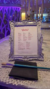 The menu for the Wonderland restaurant is displayed in a silver picture frame with filigree designs. In front of the menu sits a blue paintbrush on a black napkin, as purple lights twinkle in the background.