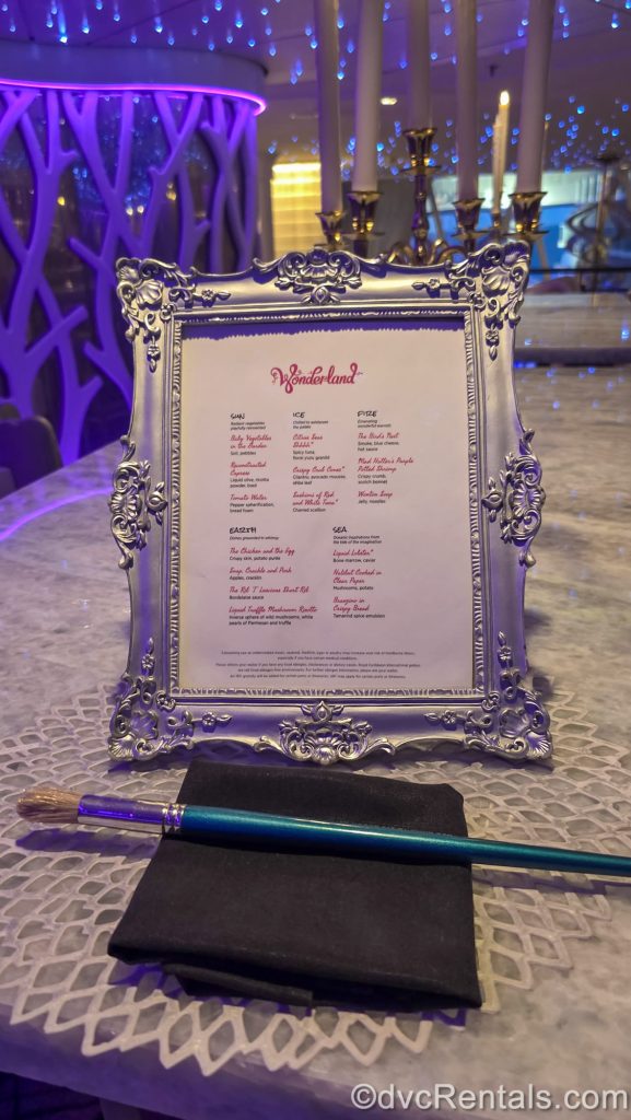 The menu for the Wonderland restaurant is displayed in a silver picture frame with filigree designs. In front of the menu sits a blue paintbrush on a black napkin, as purple lights twinkle in the background.