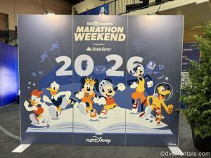 A large blue sign showing Mickey and Minnie Mouse, Goofy, Pluto, Donald and Daisy Duck running out of the pages of a storybook is displayed. The sign reads “Walt Disney World Marathon Weekend 2026” on it in white lettering.