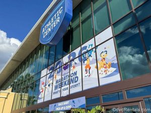 The Athletic Center at the ESPN Wide World of Sports Complex is seen under a sunny sky. Above the entrance is blue and white Marathon Weekend signage indicating race shirt pick-up and expo are inside.
