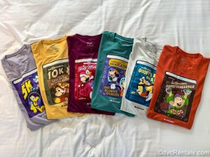 Six multicolored shirts each themed to a different runDisney Marathon event are folded and laid out on display. Each shirt has images of different Disney characters and storybook-theming.