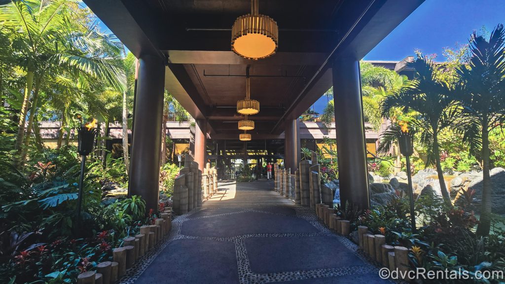 A covered grey stone and wood pathway lined with tropical landscaping, tiki torches and water features leads to the entrance of Disney’s Polynesian Villas & Bungalows at Walt Disney World.