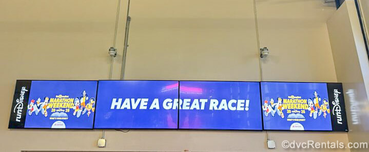 Monitors displaying the runDisney Walt Disney World Marathon Weekend logo and the words “Have A Great Race!” in white writing on a blue background are seen.
