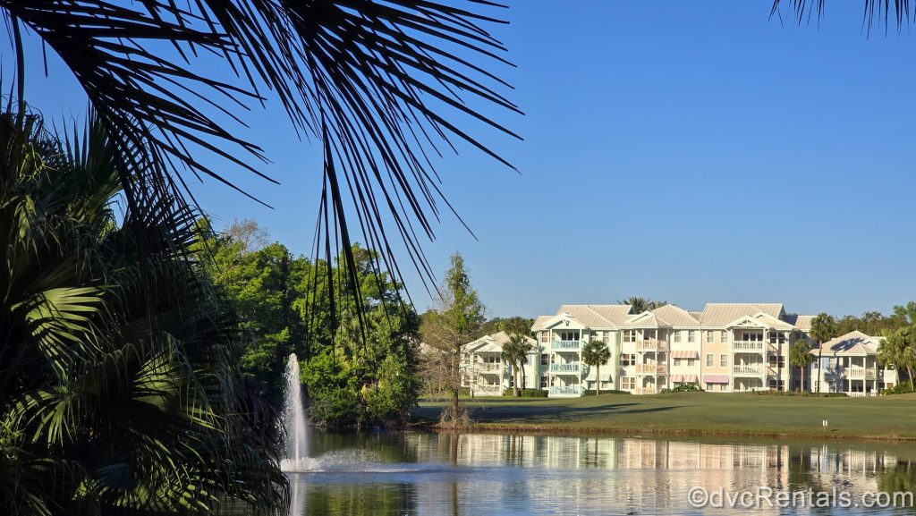 Greenery, water, a fountain, and the pastel colored villa buildings are seen under a bright blue sky at Disney’s Old Key West Resort.
