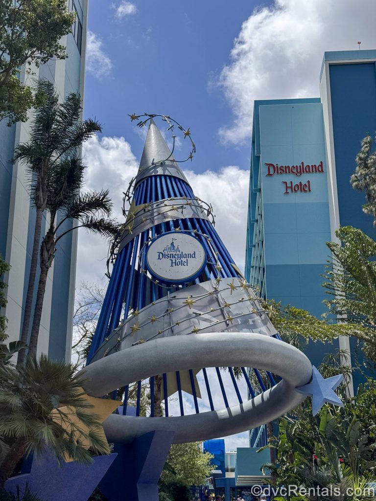 A larger than life blue and silver wizard hat wrapped in gold stars displays a white circular sign for the Disneyland Hotel in Anaheim, California, with the resort exterior visible in the background.