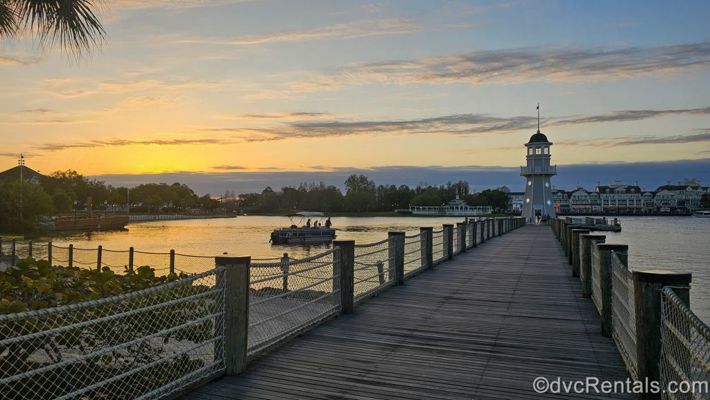 The sunrise paints the sky over Disney’s BoardWalk and Beach Club Villas with shades of blue, yellow, and orange as a small boat sails over Cresent Lake.