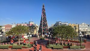 A tall Christmas tree decorated with red bows is displayed at the Magic Kingdom under a sunny blue sky. Other Christmas decorations such as nutcrackers and garlands are seen on Main Street U.S.A. as well.