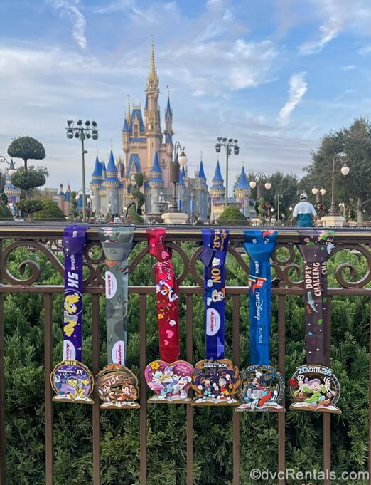 Six different runDisney medals featuring different Disney characters are displayed on a fence in front of Cinderella Castle in the Magic Kingdom at Walt Disney World under a blue sky.