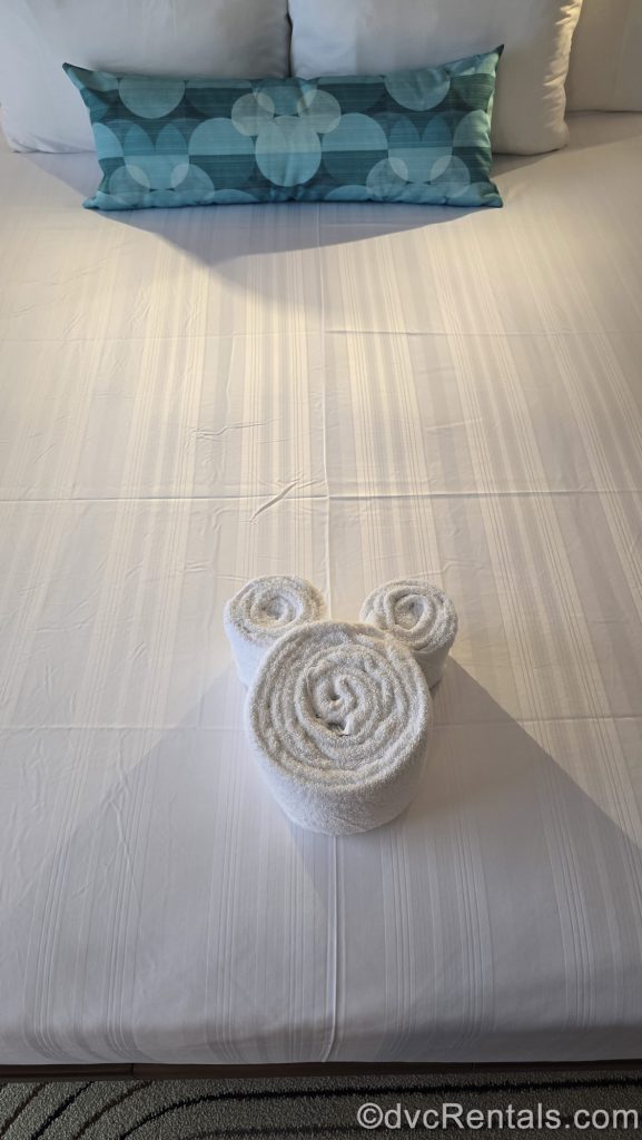 A white towel rolled to make the shape of Mickey Mouse’s head is displayed on a bed along with a turquoise throw pillow featuring Mickey at Bay Lake Tower at Disney’s Contemporary Resort.