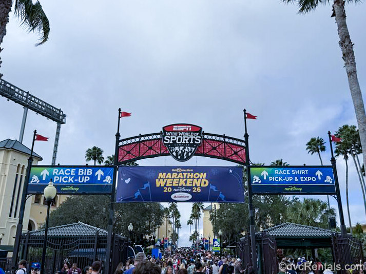 Crowds of runDisney participants enter the ESPN Wide World of Sports Complex at Walt Disney World under colorful signage indicating Marathon Weekend race bib and shirt pick-up locations ahead.