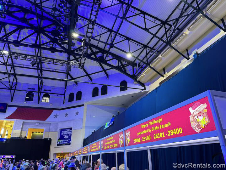 Marathon Weekend runDisney participants line up to pick up their race bibs at the ESPN Wide World of Sports Complex. There is red signage indicating each race at the top of each booth.