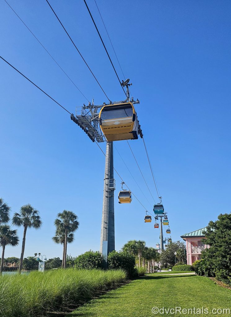 Colorful Disney Skyliner gondolas travel along their cables through a blue sky with resort buildings and greenery below.