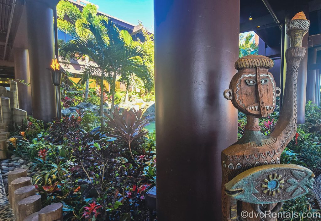 A carved Tiki statue is seen outside the entryway to Disney’s Polynesian Villas & Bungalows. There is tropical foliage in the background as well as a torch lit with fire and a stone pathway leading into the resort.