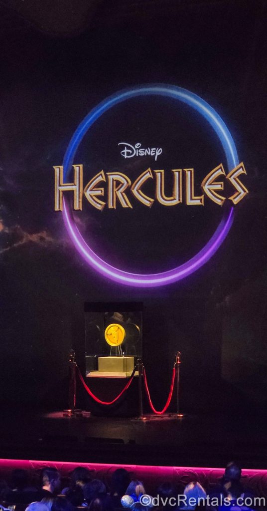 “Disney Hercules” is seen displayed in white and gold writing in the middle of a pink and blue circle above a display of a golden coin on stage ahead of the stage production on the Disney Destiny.